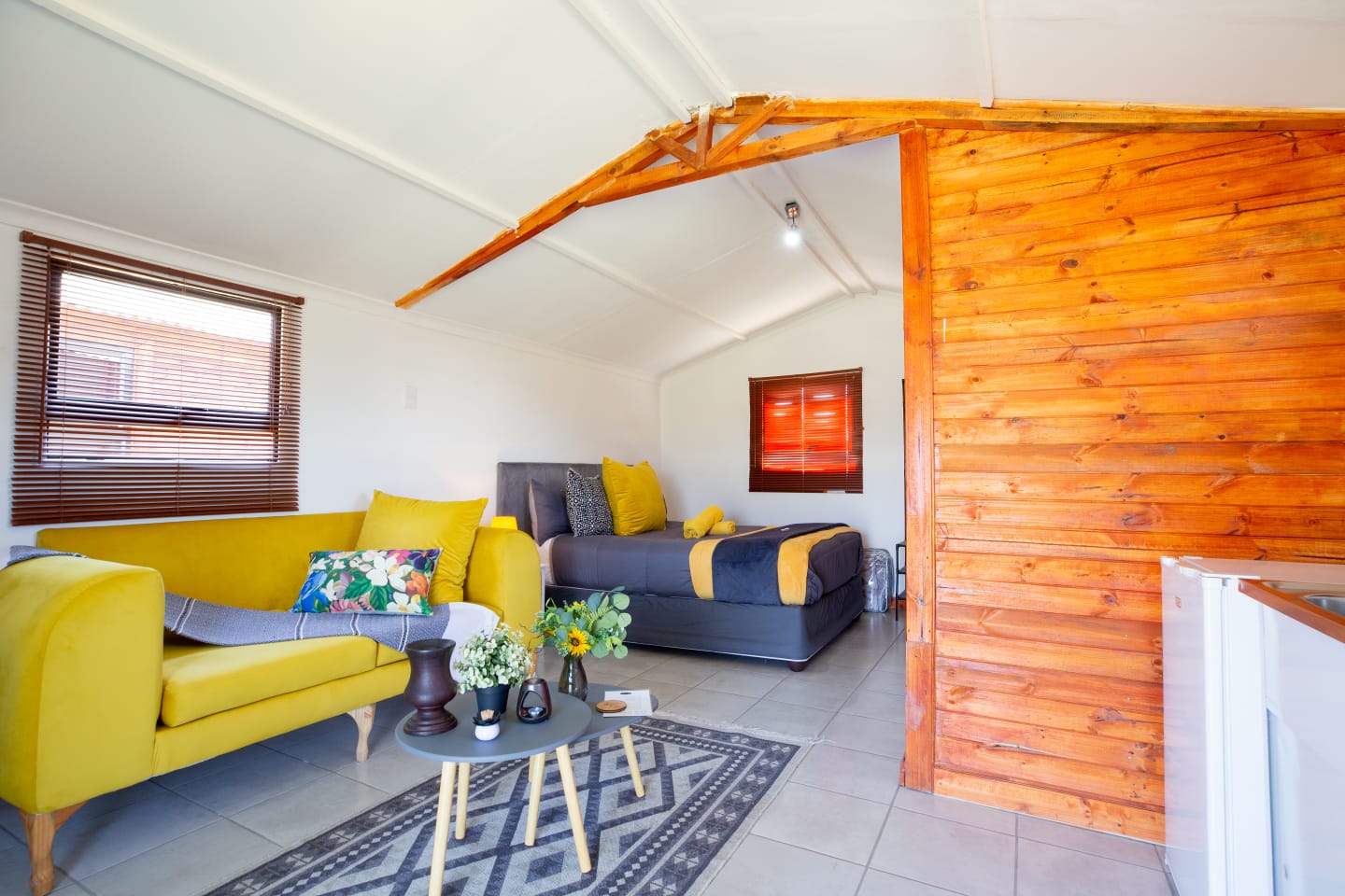 A bright open cabin interior with a lounge, bed, kitchenette, wooden feature wall, and cheerful yellow accents.